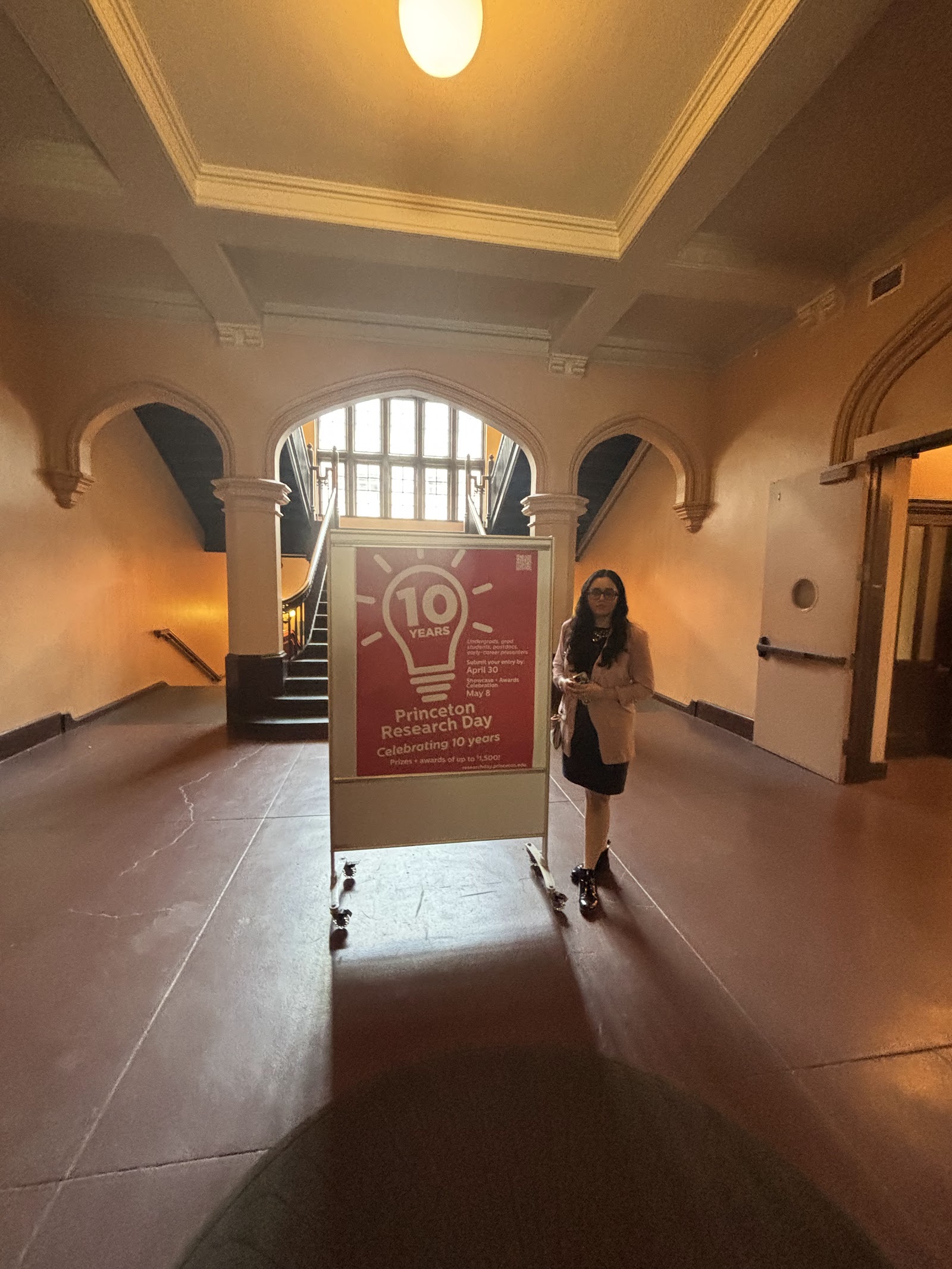 Shalvi standing beside a large sign reading “10 — Princeton Research Day, Celebrating 10 years” in an arched stone hallway.