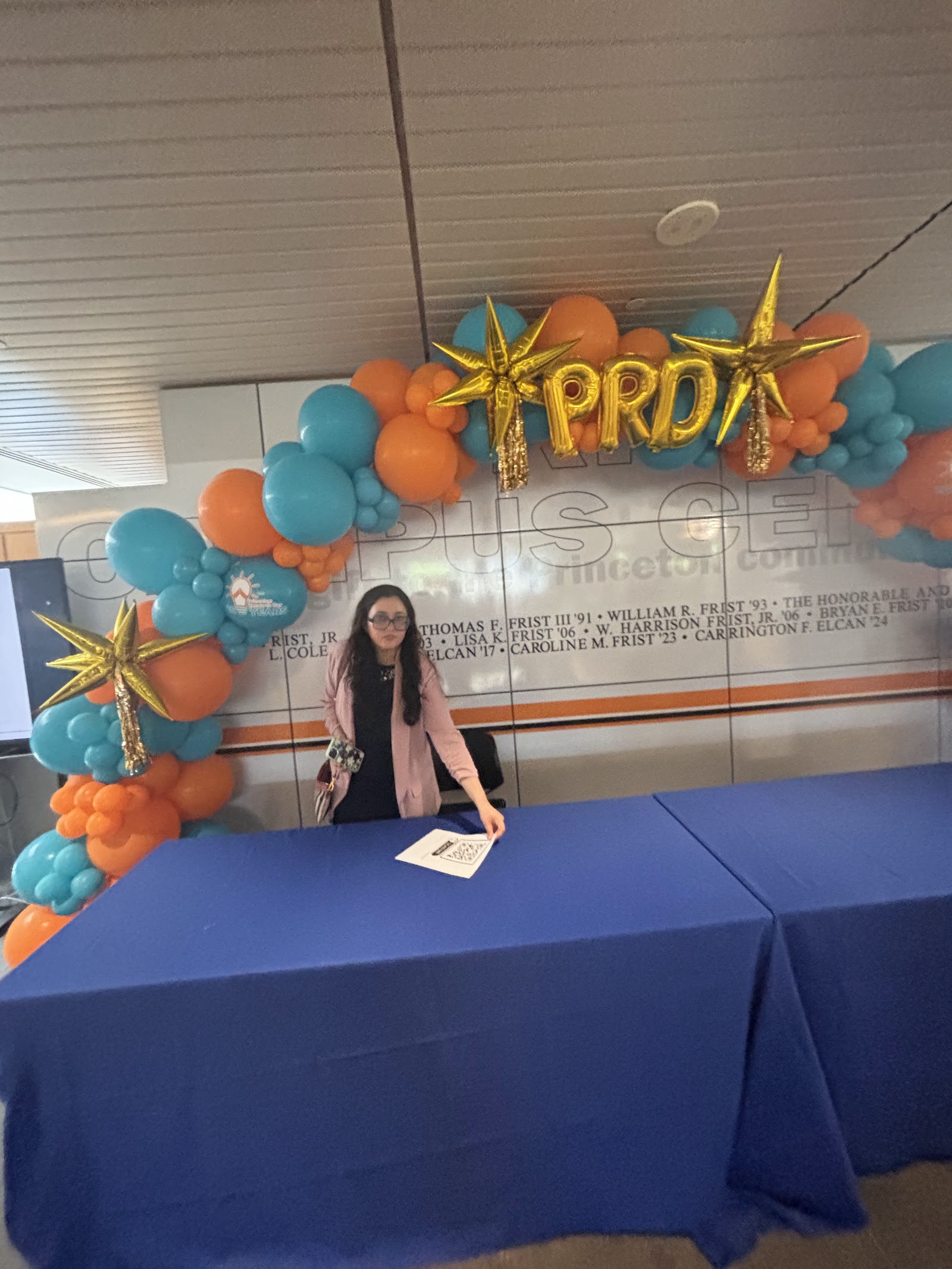 Shalvi at a blue-draped table under a balloon arch with gold stars spelling PRD.