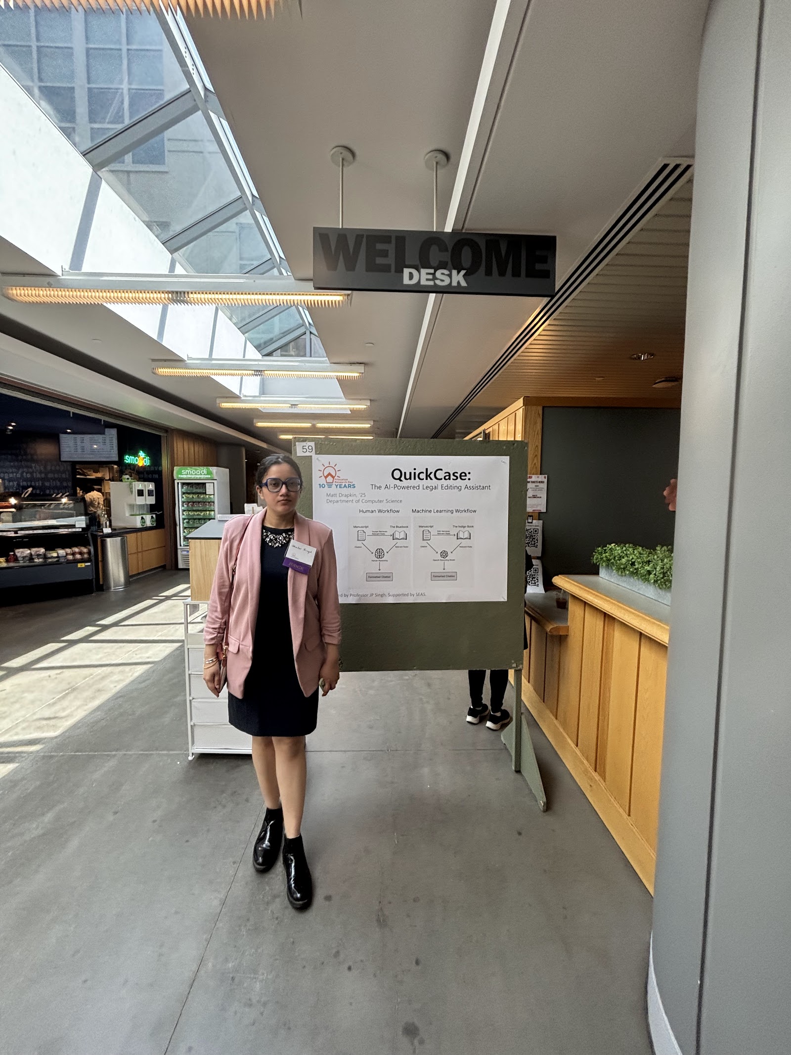 Shalvi under a “Welcome Desk” sign beside a QuickCase poster in a skylit atrium.