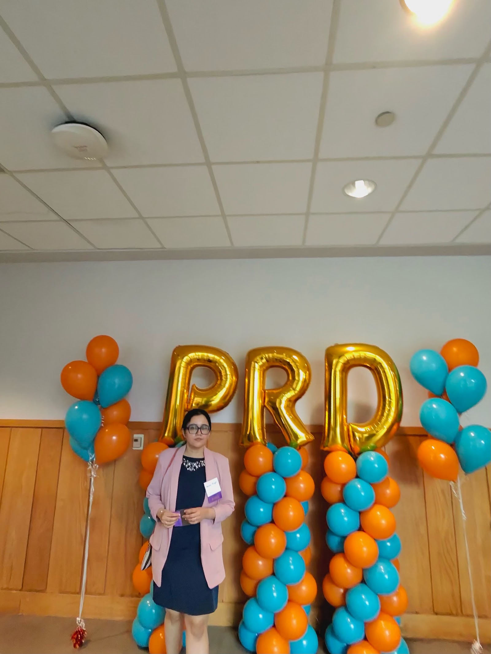 Shalvi in front of giant gold foil balloon letters spelling PRD, flanked by orange and teal balloon columns.
