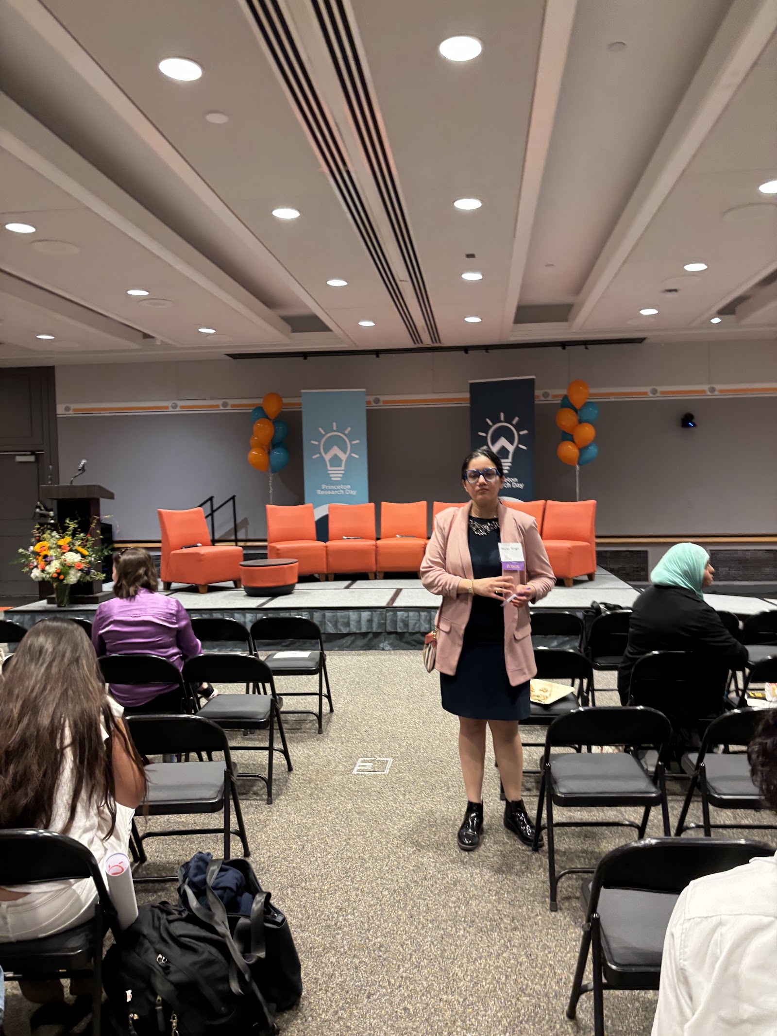 Shalvi standing in the ballroom before an empty stage set with orange couches.
