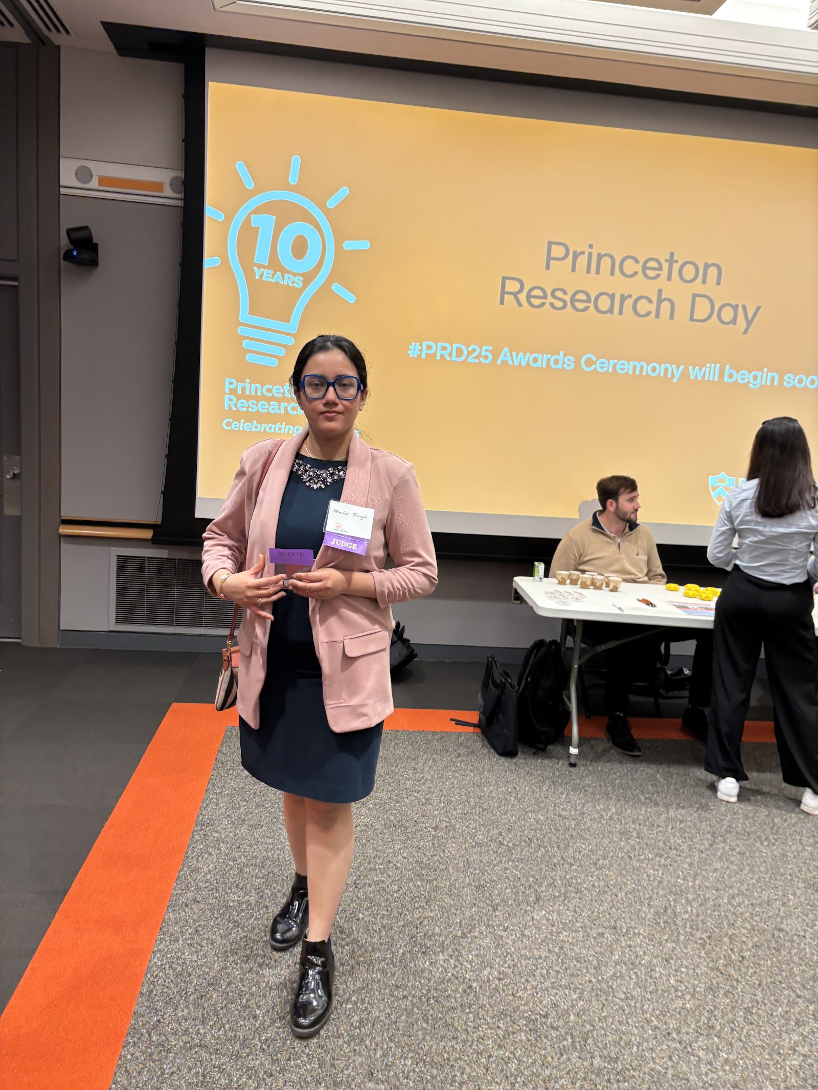 Shalvi holding her badge in front of a screen reading “Princeton Research Day — #PRD25 Awards Ceremony will begin soon.”
