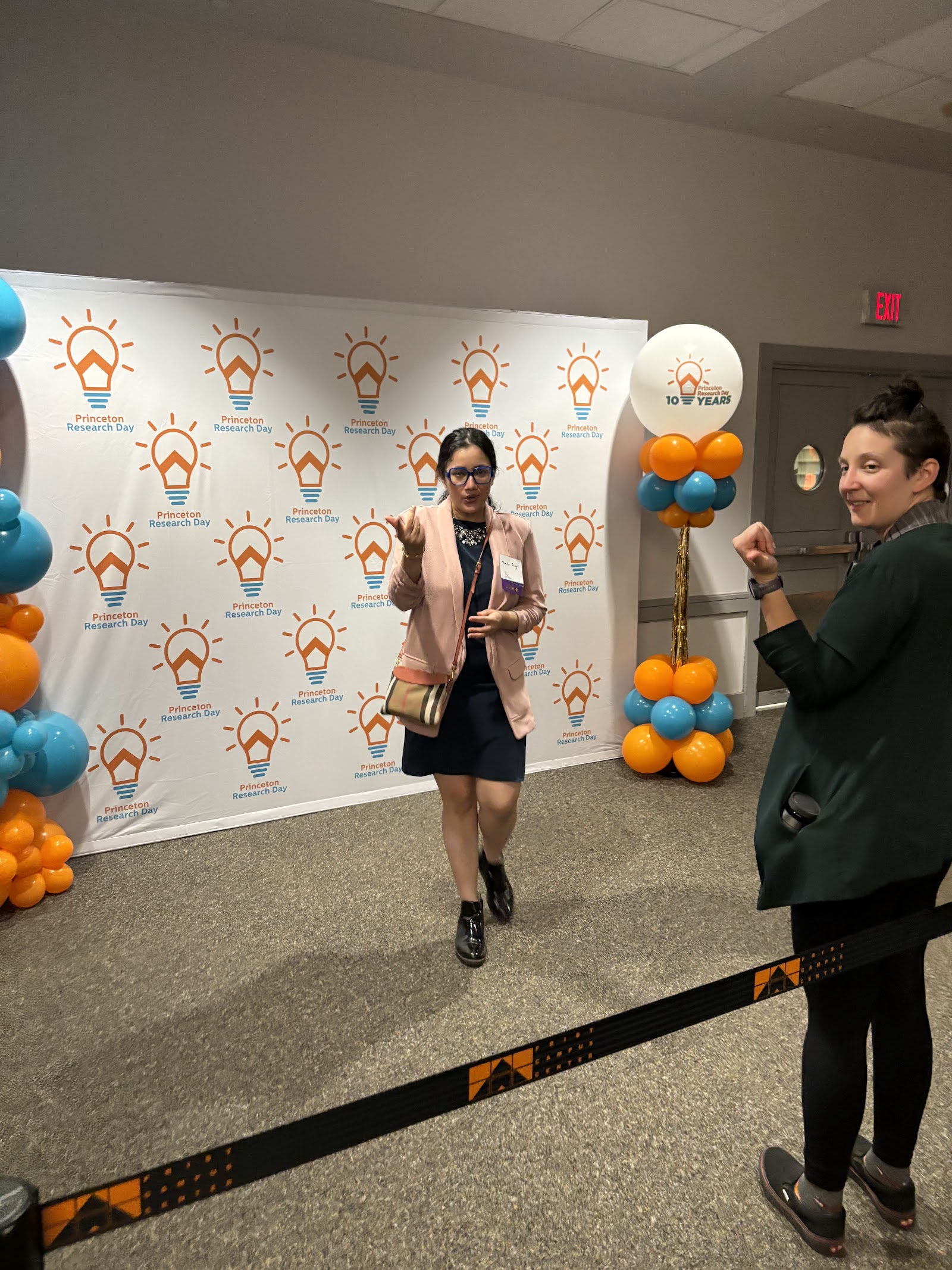 Shalvi posing on a PRD-branded step-and-repeat backdrop while someone cheers her on.