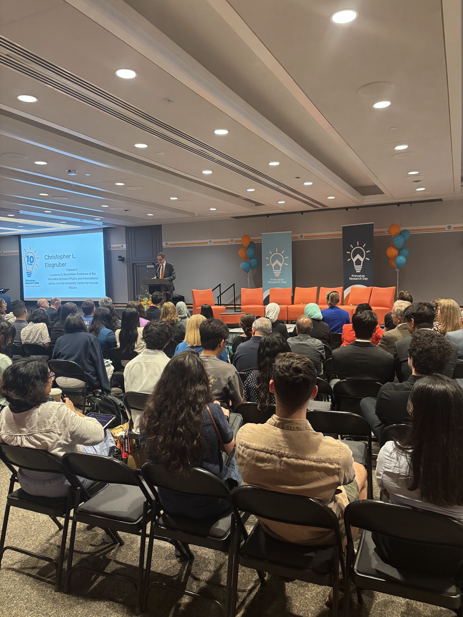 A full audience watching a speaker on stage under the Research Day screen.