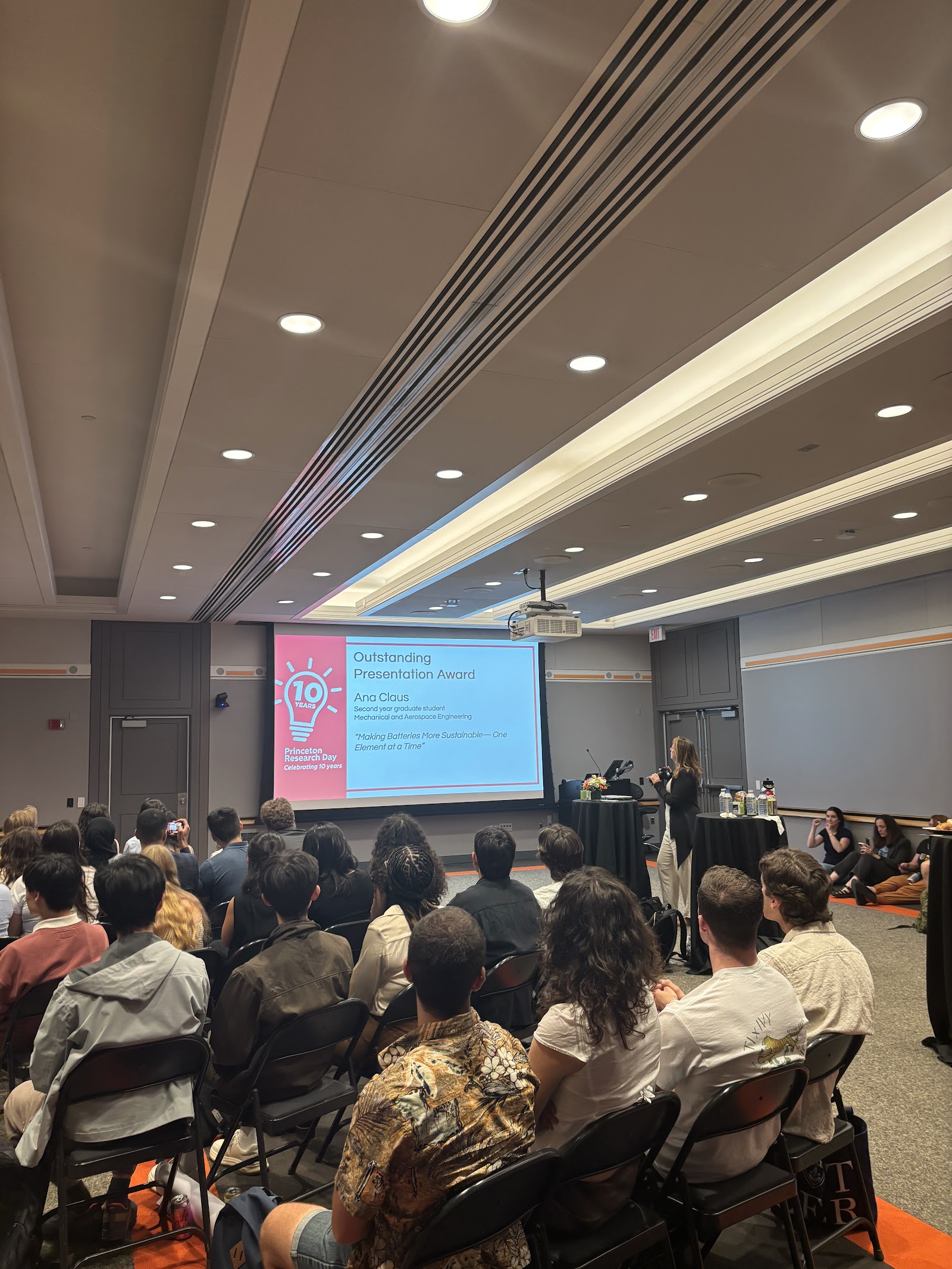 A screen reading “Outstanding Presentation Award” in front of a seated crowd.