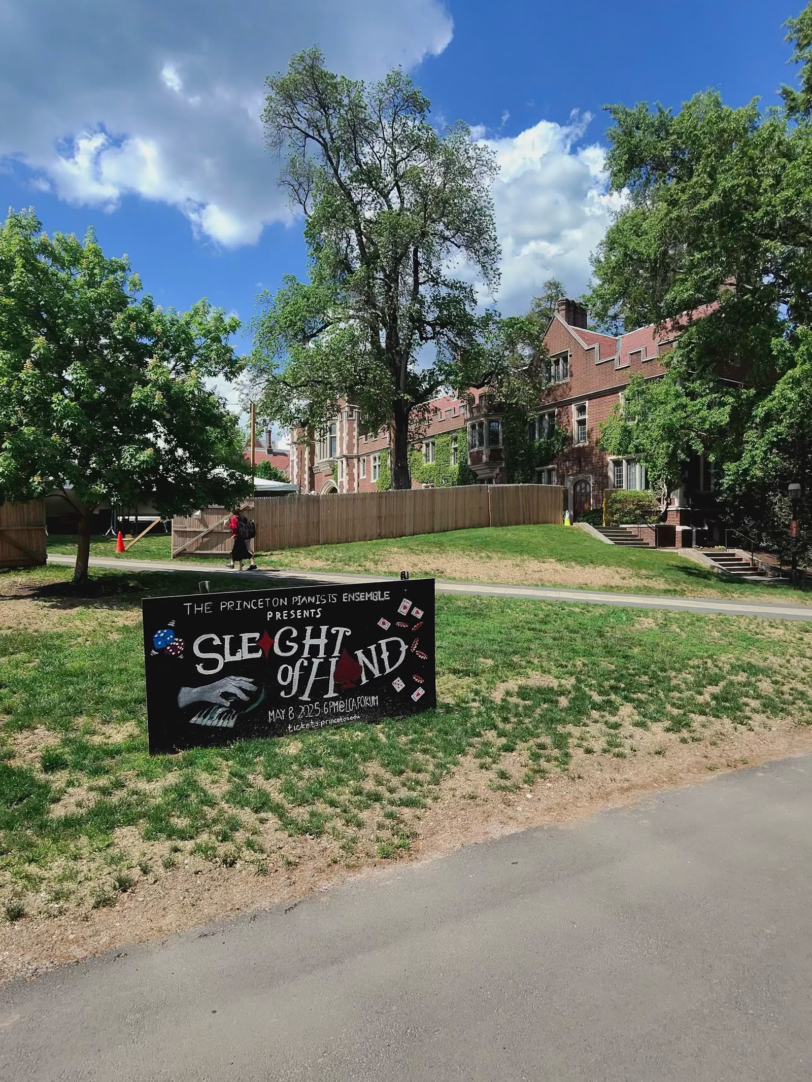 A hand-lettered chalkboard sign reading “Sleight of Hand” on a lawn in front of a brick building.